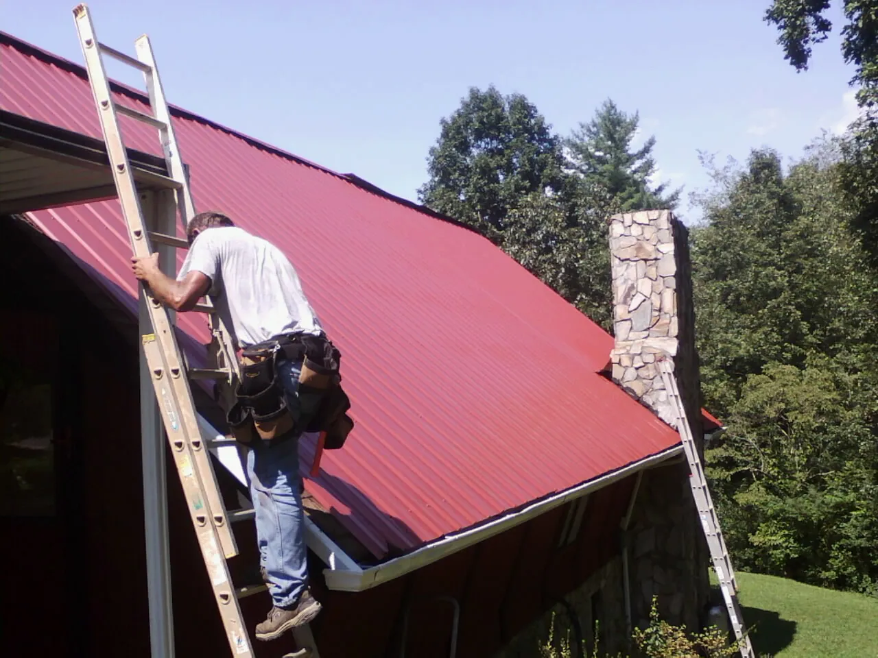 Roofer on a ladder beside a red metal roof and stone chimney — Soffit & Fascia Repair by Roofing Brothers in Oswego